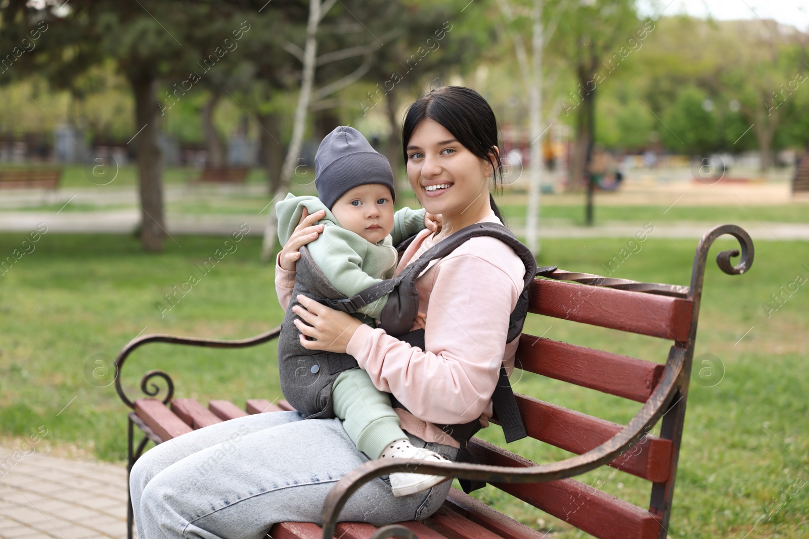 Photo of Mother holding her child in sling (baby carrier) on bench in park