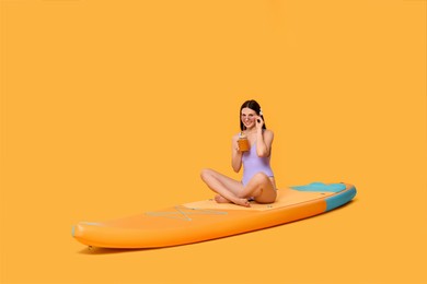 Photo of Happy woman with refreshing drink resting on SUP board against orange background