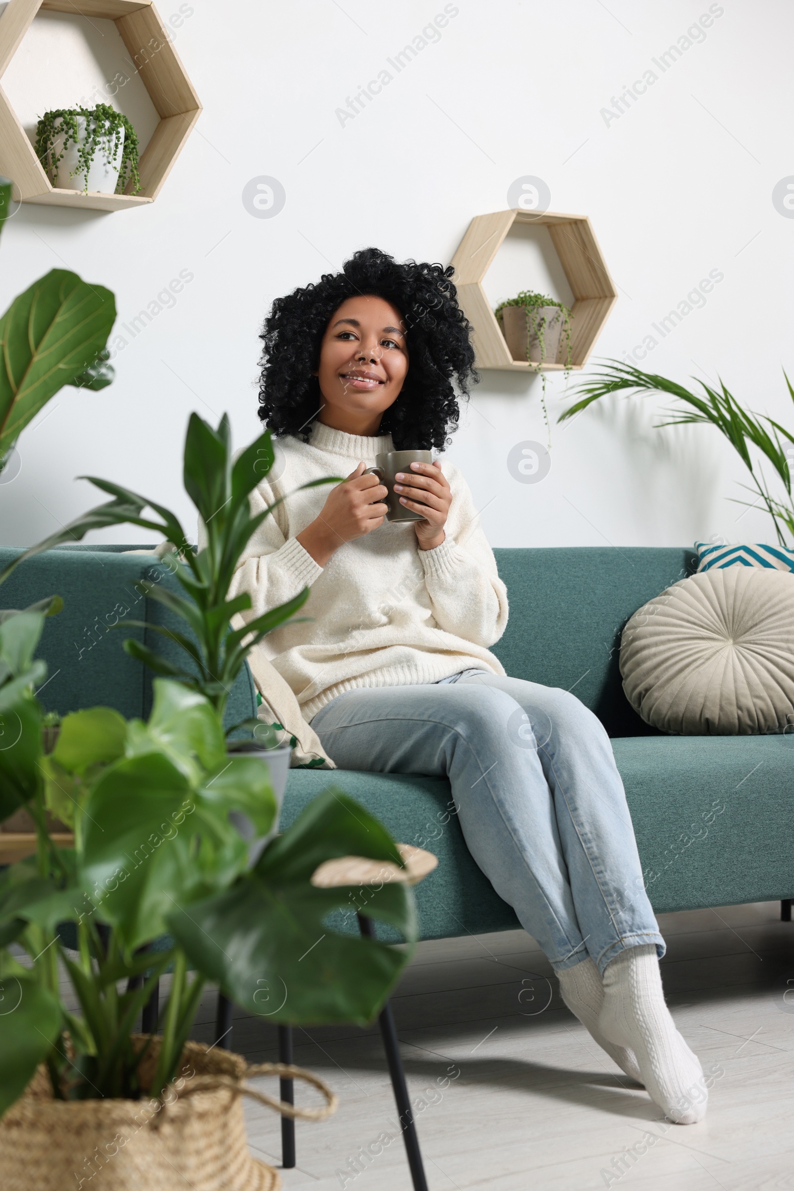 Photo of Relaxing atmosphere. Happy woman with cup of hot drink on sofa near beautiful houseplants in room