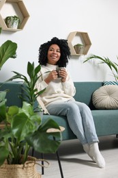 Relaxing atmosphere. Happy woman with cup of hot drink on sofa near beautiful houseplants in room