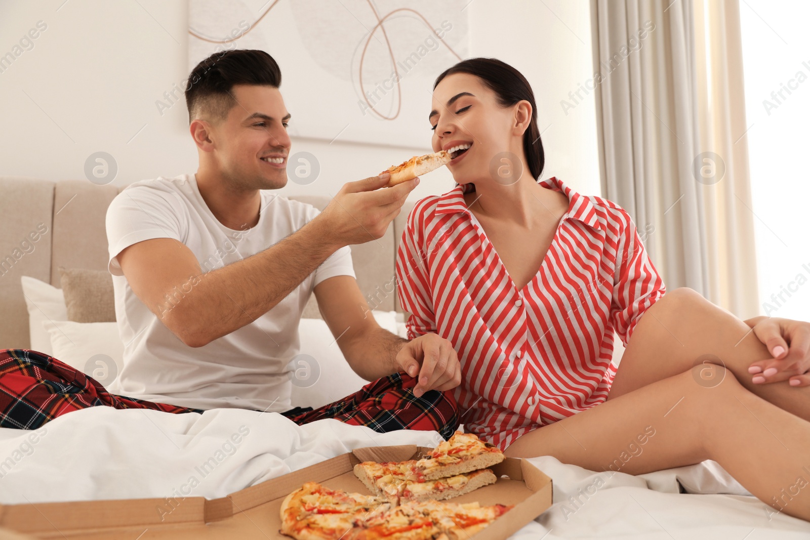 Photo of Happy couple in pyjamas eating pizza on bed at home