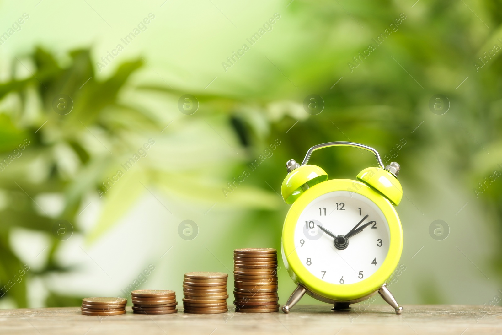 Photo of Light green alarm clock and stacked coins on wooden table against blurred background. Money savings