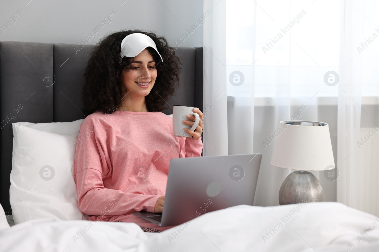 Photo of Beautiful young woman in stylish pyjama and sleep mask with cup of drink using laptop in bed at home