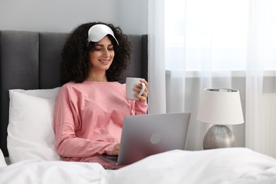 Photo of Beautiful young woman in stylish pyjama and sleep mask with cup of drink using laptop in bed at home