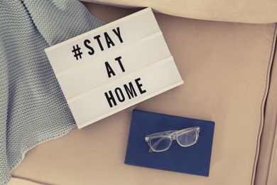 Photo of Book, glasses, plaid and lightbox with hashtag STAY AT HOME on sofa, flat lay. Message to promote self-isolation during COVID‑19 pandemic
