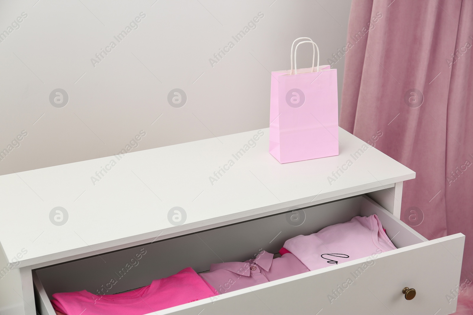 Photo of White chest of drawers with pink clothes and bag indoors