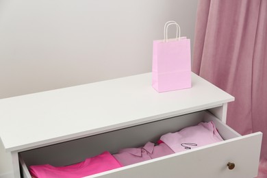 Photo of White chest of drawers with pink clothes and bag indoors
