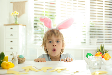 Emotional little boy wearing bunny ears headband at table with Easter eggs, indoors