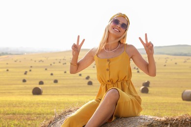 Photo of Beautiful hippie woman showing peace signs on hay bale in field, space for text