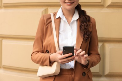 Woman in stylish suit using smartphone near pale yellow wall outdoors, closeup