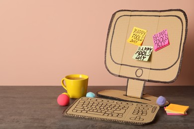 Photo of Cardboard computer with Happy Fools' Day notes and cup on wooden table. Space for text