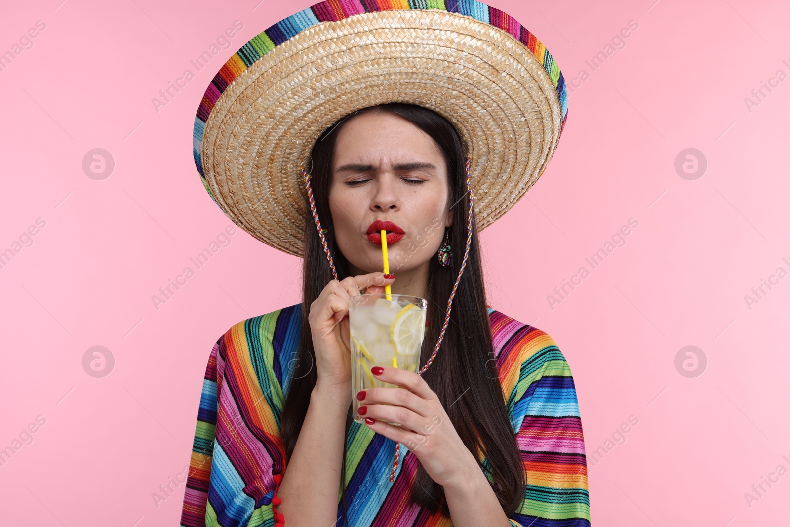 Photo of Young woman in Mexican sombrero hat and poncho drinking cocktail on pink background