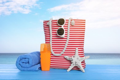 Image of Stylish striped bag and other beach accessories on light blue wooden table near sea