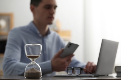 Photo of Hourglass with flowing sand on desk. Man using smartphone while working on laptop indoors, selective focus