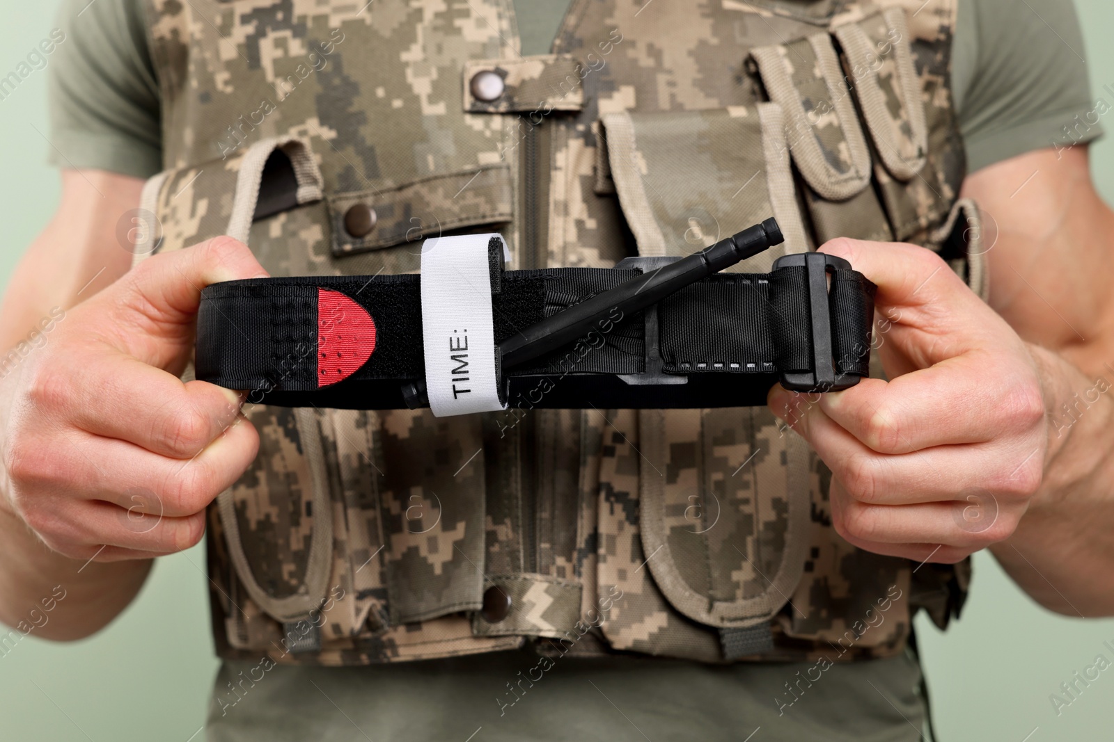 Photo of Soldier in military uniform holding medical tourniquet on light olive background, closeup