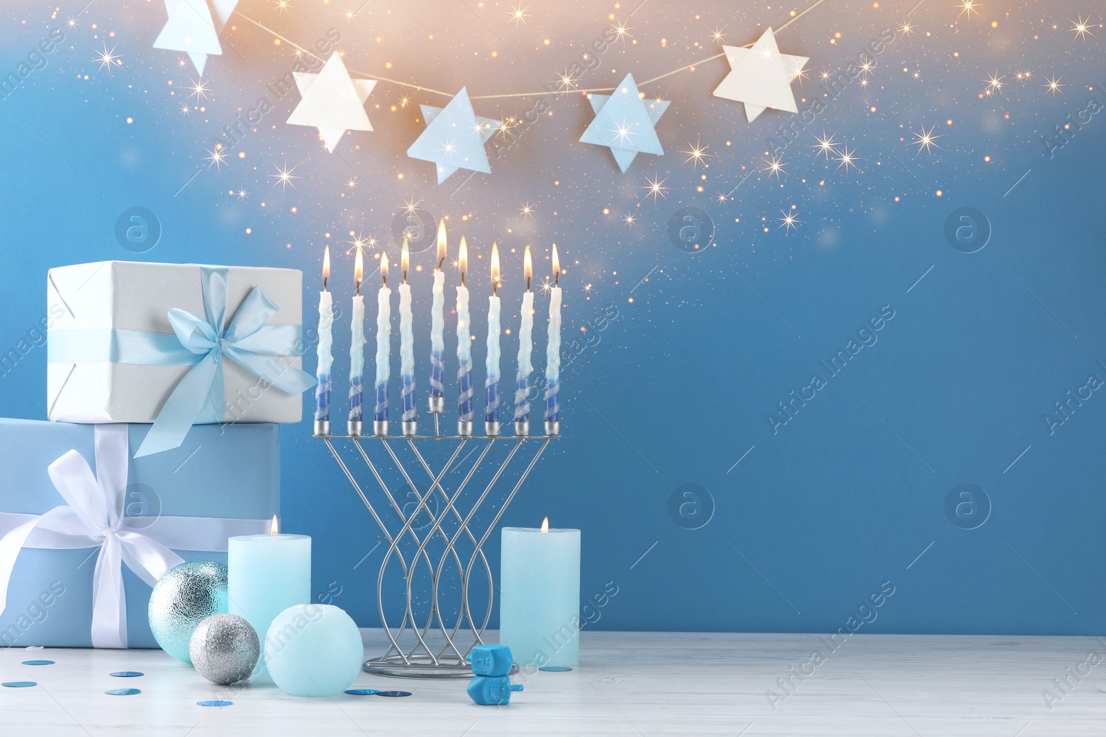 Image of Hanukkah celebration. Menorah, burning candles, dreidels and gift boxes on white wooden table against light blue background, space for text