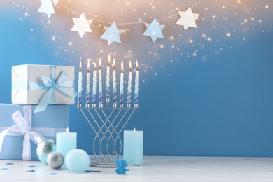 Image of Hanukkah celebration. Menorah, burning candles, dreidels and gift boxes on white wooden table against light blue background, space for text