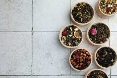 Different kinds of dry herbal tea in wooden bowls on white tiled surface, flat lay. Space for text