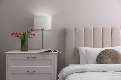 Photo of Stylish lamp, flowers and magazine on bedside table indoors. Bedroom interior elements