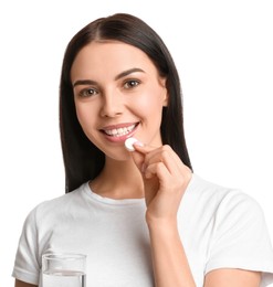 Young woman with glass of water taking vitamin pill on white background