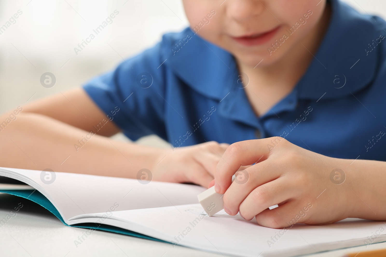 Photo of Little boy erasing mistake in his notebook at white desk, closeup