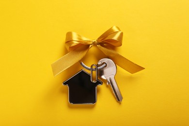 Photo of Key with trinket in shape of house and bow on yellow background, top view. Housewarming party
