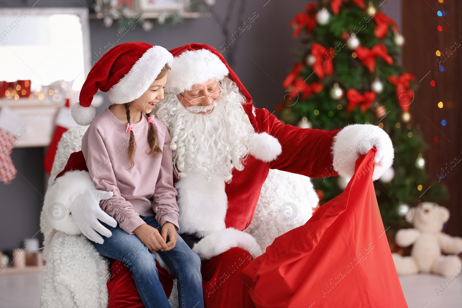 Photo of Authentic Santa Claus showing his bag with gifts to little girl indoors