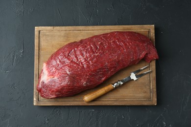 Photo of Wooden board with piece of raw meat and fork on black table, top view