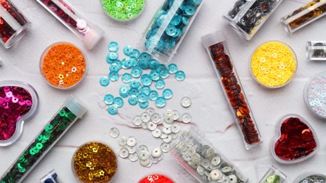 Photo of Many different colorful sequins in containers and tubes on marble e background, flat lay