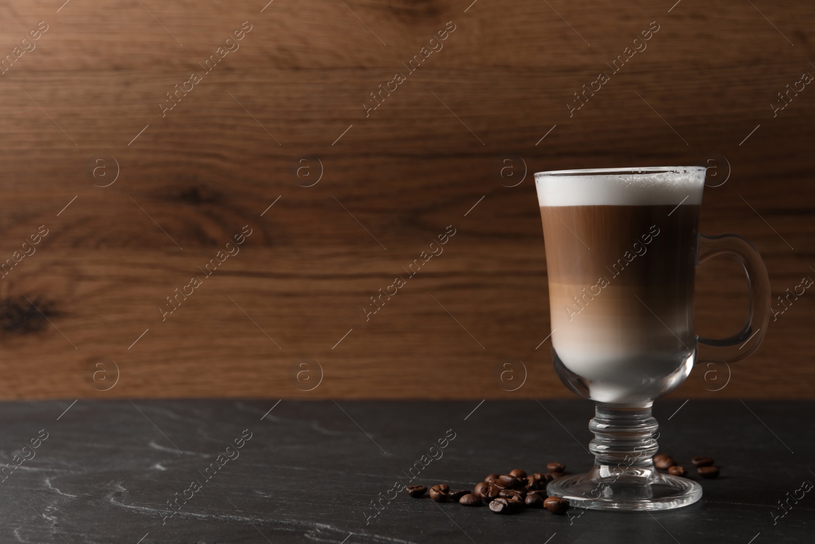 Photo of Glass cup of delicious layered coffee and beans on black table against wooden background, space for text