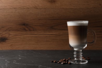 Glass cup of delicious layered coffee and beans on black table against wooden background, space for text