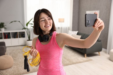 Happy sports blogger holding skipping rope while streaming online fitness lesson with smartphone at home