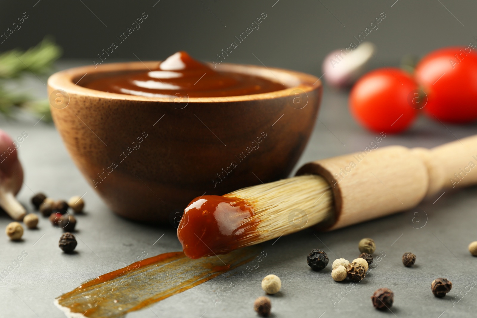 Photo of Tasty barbeque sauce in bowl, brush and peppercorns on grey table, closeup