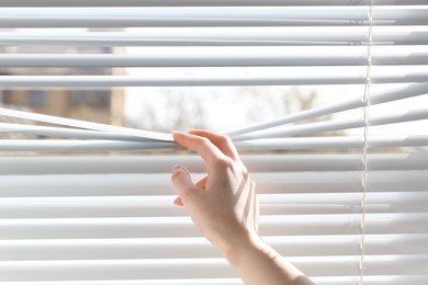 Photo of Woman separating slats of white blinds indoors, closeup