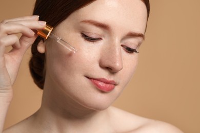 Beautiful woman with freckles applying cosmetic serum onto her face on beige background, closeup