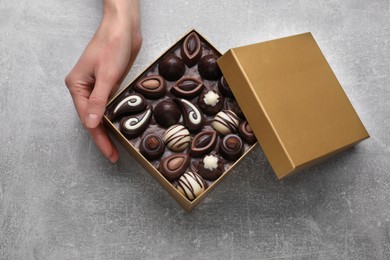 Photo of Woman with open box of delicious chocolate candies at light grey table, top view