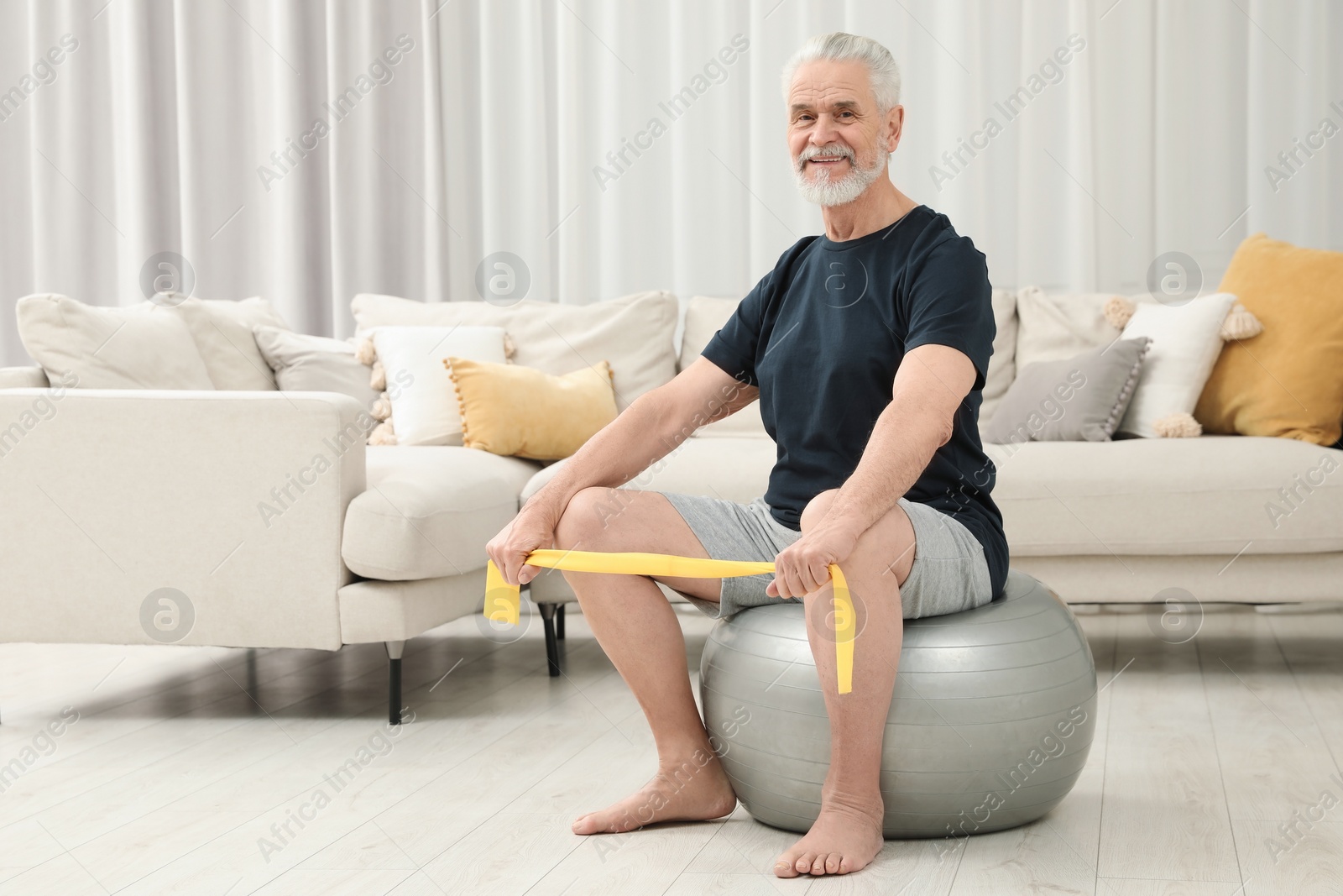 Photo of Senior man doing exercise with elastic resistance band on fitness ball at home