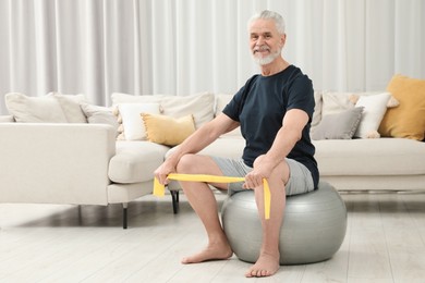 Senior man doing exercise with elastic resistance band on fitness ball at home