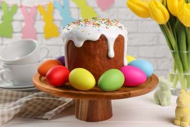 Photo of Stand with tasty Easter cake and decorated eggs on white wooden table