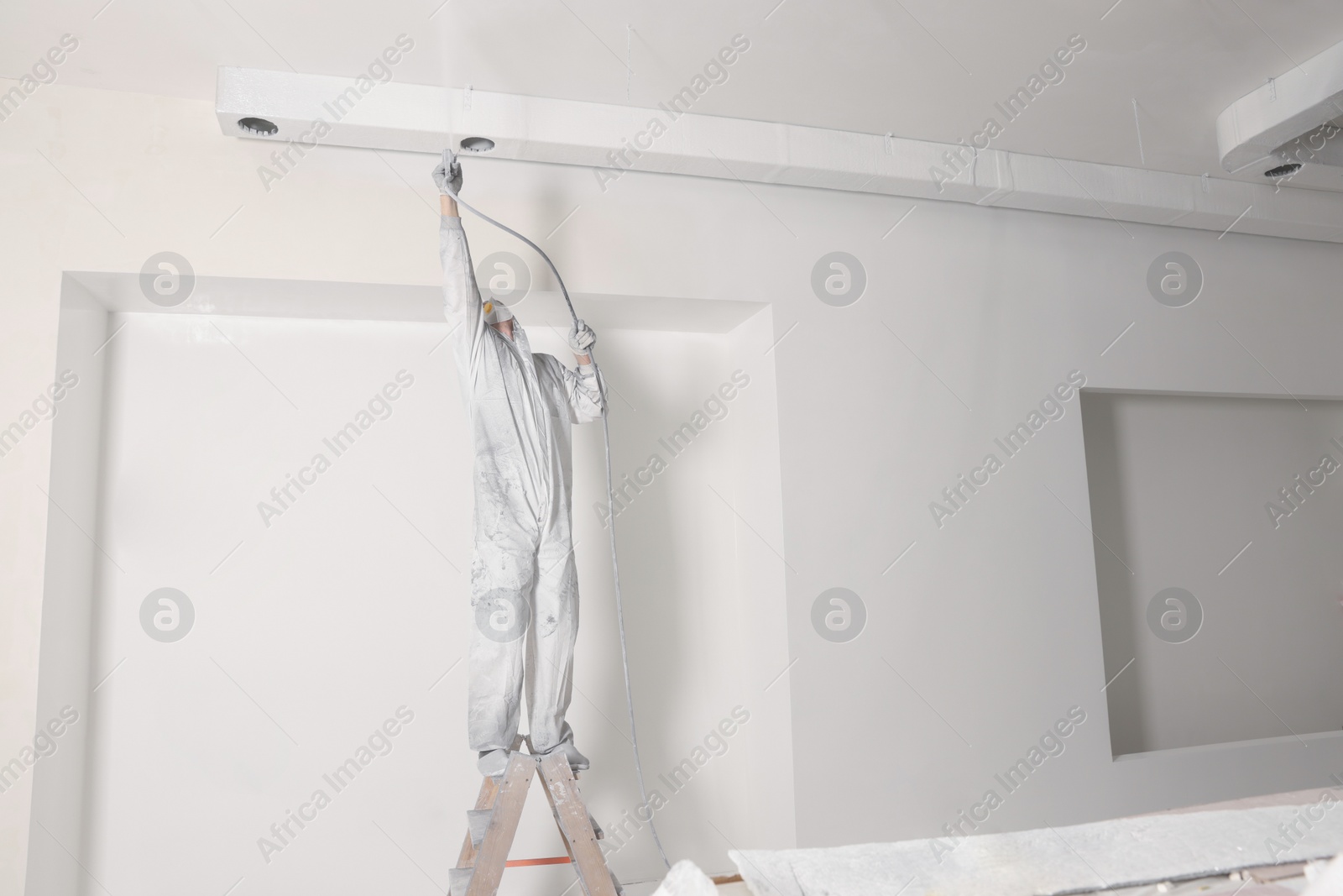 Photo of Decorator in protective overalls painting wall with spray gun indoors