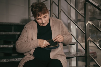 Poor senior woman with empty wallet and coin sitting on stairs indoors