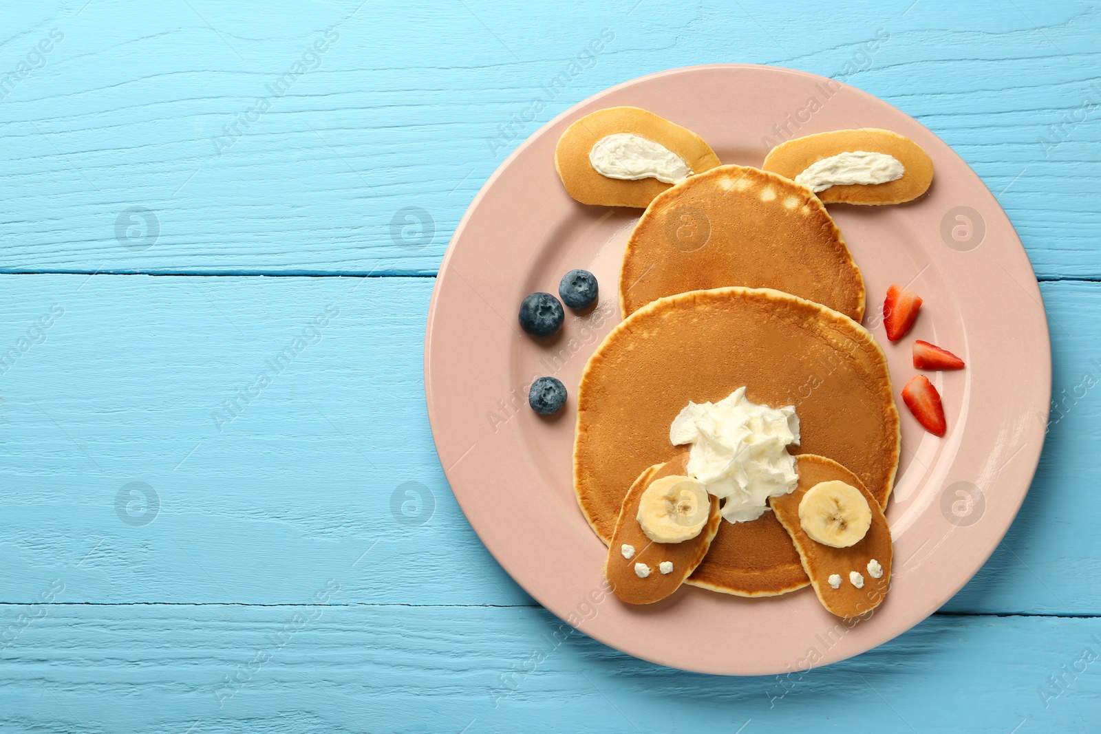 Photo of Creative serving for kids. Plate with cute bunny made of pancakes, berries, cream and banana on light blue wooden table, top view. Space for text