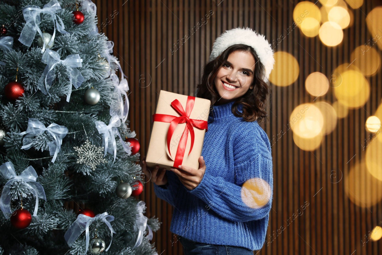 Image of Happy young woman in Santa hat with gift near Christmas tree at home, bokeh effect