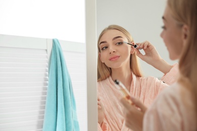 Photo of Young woman applying oil onto her eyelashes near mirror indoors