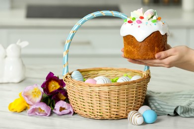 Photo of Woman holding tasty Easter cake near wicker basket with painted eggs and flowers at white marble table, closeup