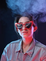 Young woman wearing sunglasses in smoke on color background