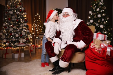Little girl whispering in Santa Claus' ear near Christmas tree indoors