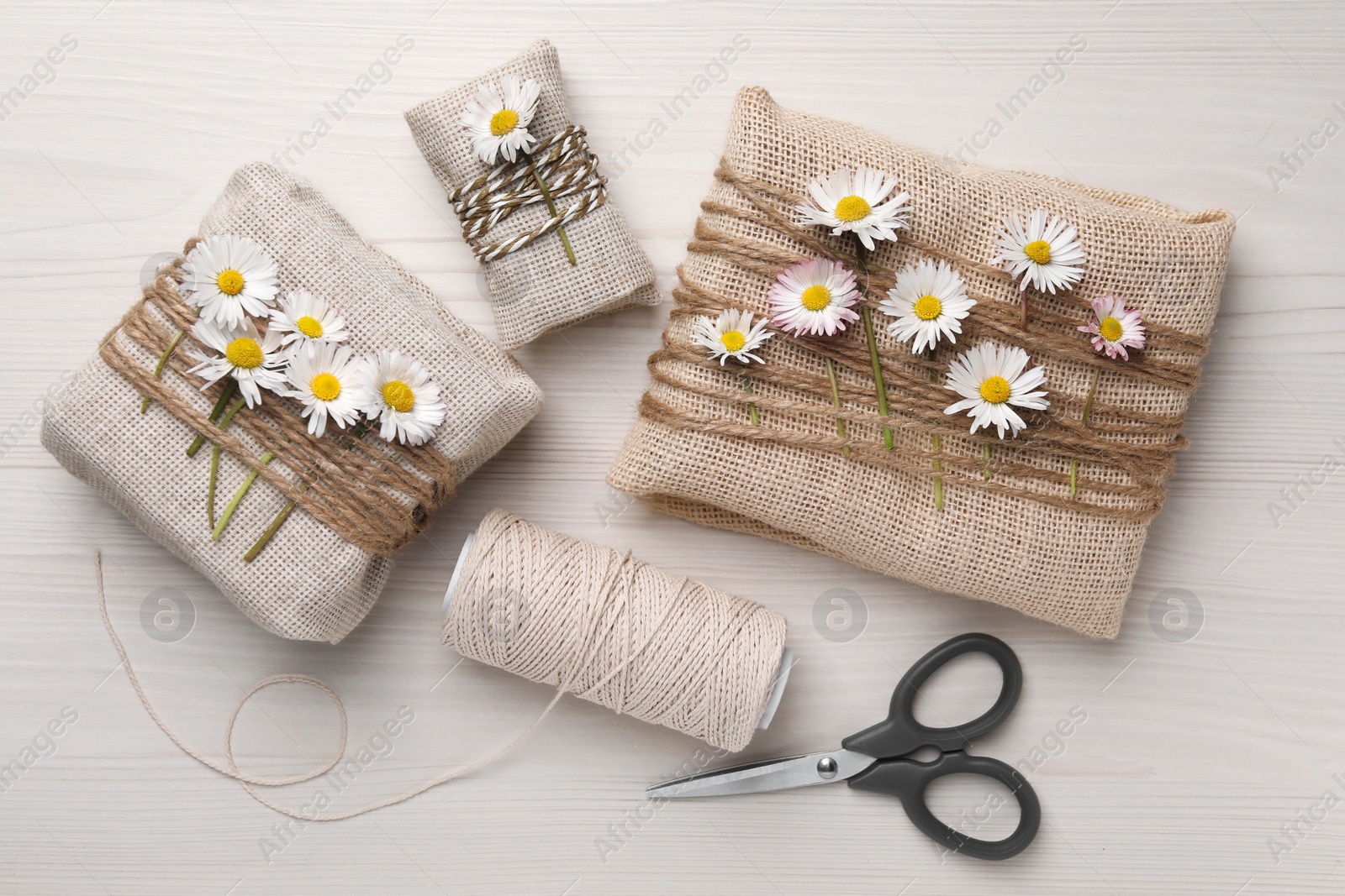 Photo of Gifts packed in burlap fabric with beautiful chamomiles, ropes, spool of threads and scissors on white wooden table, flat lay