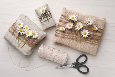 Photo of Gifts packed in burlap fabric with beautiful chamomiles, ropes, spool of threads and scissors on white wooden table, flat lay