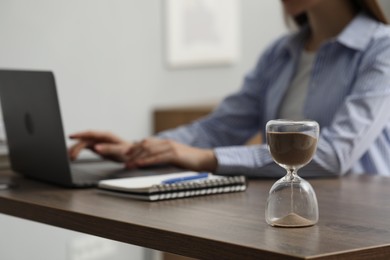 Hourglass with flowing sand on desk. Woman using laptop indoors, selective focus